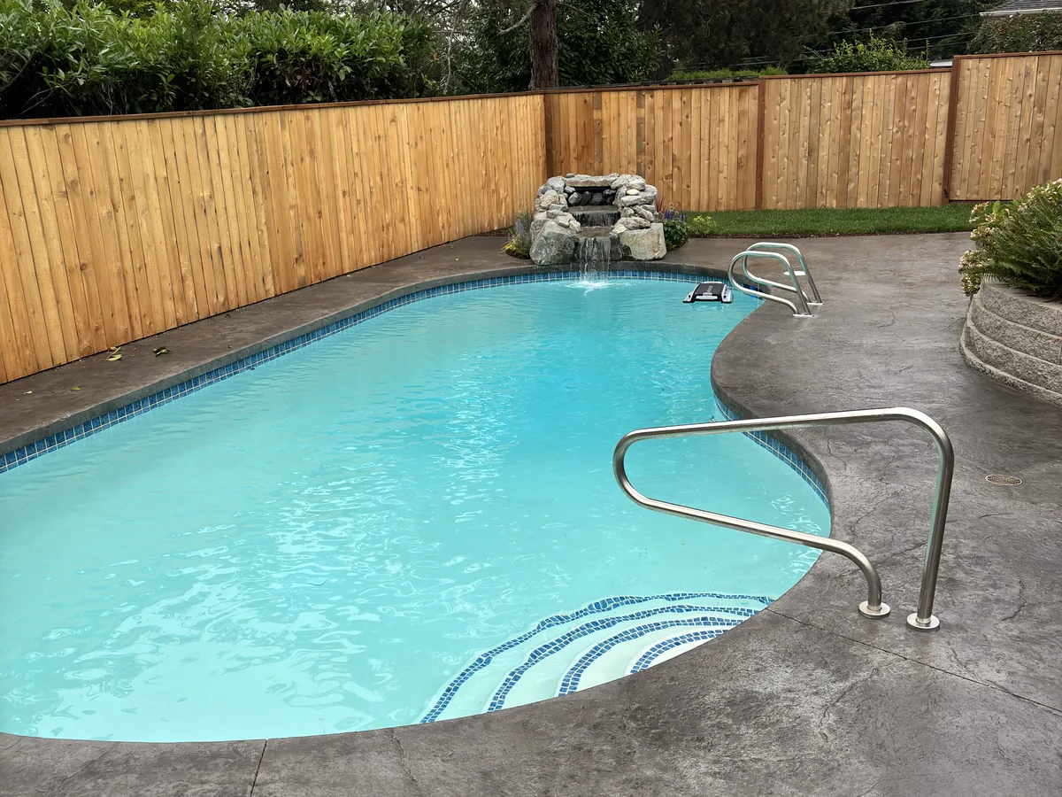 Finished pool renovation with new tile, stamped concrete deck, cedar privacy fence, and rock waterfall