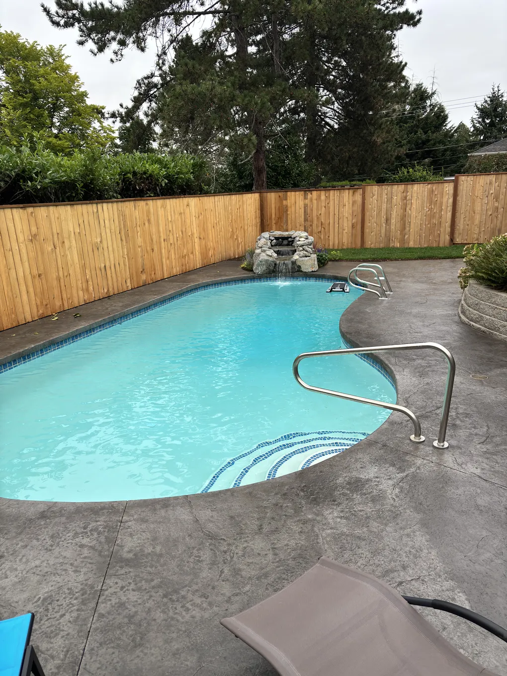 Finished pool renovation with new tile, stamped concrete deck, cedar privacy fence, and rock waterfall